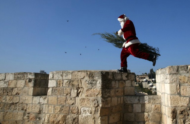 Un palestino cristiano vestido como Papá Noel camina llevando un árbol de Navidad a lo largo de las paredes viejas de la ciudad de Jerusalén el 21 de diciembre de 2015, como los cristianos de todo el mundo se preparan para celebrar la Navidad. AFP PHOTO / GALI Tibbon