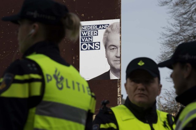 Dutch police officers stand guard next to a campaign poster of Dutch far-right Freedom Party leader (Partij Voor De Vrijheid - PVV) Geert Wilders as he meets with supporters in Heerlen on March 11, 2017. Wilders was guarded by police as protesters took to the streets while he handed out pamphlets and posed for selfies as part of his election campaign. / AFP PHOTO / JOHN THYS